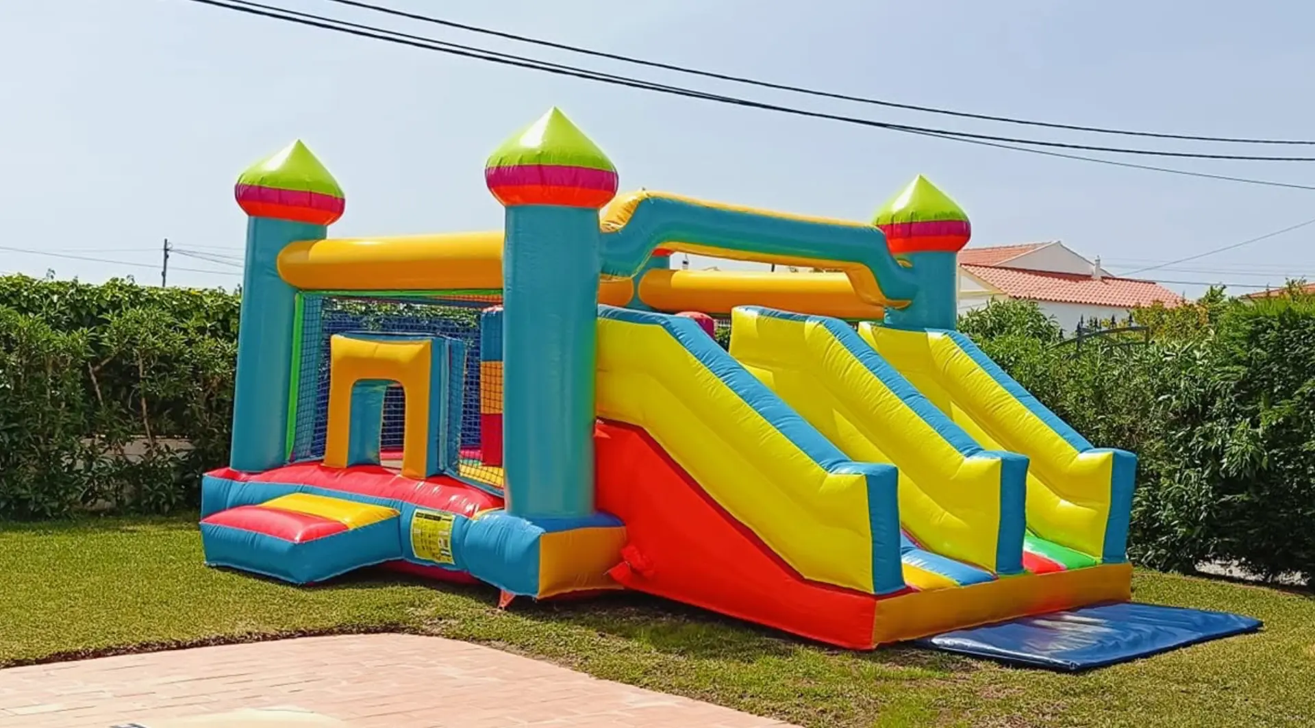 Insuflável Brincar em forma de castelo com escorrega duplo, torres com pontas cónicas verdes e rosa em tons de azul, amarelo e vermelho, montado num relvado