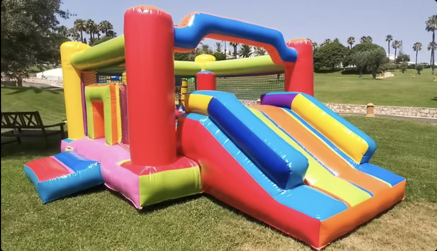 Insuflável Kids com escorrega duplo frontal em faixas amarelas, azuis e laranja, pilares vermelhos e amarelos, arco de entrada verde e laranja e obstáculos interiores, montado num relvado